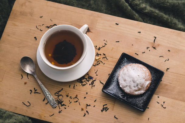 Homemade cupcake with powdered sugar on a black plate and a white cup of tea with natural additives on a wooden tray on a background of green textiles. The concept of a cozy breakfast in bed - Foto, immagini