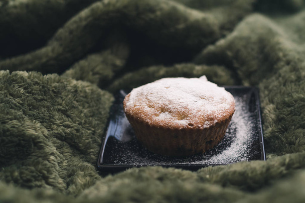 Homemade cupcake with powdered sugar on a black plate on a background of green textiles. The concept of a cozy breakfast in bed - Fotografie, Obrázek
