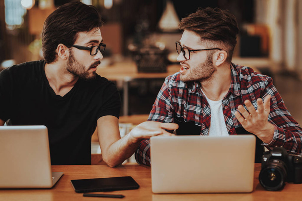 Two Freelance Photographers Work on Laptops. Portrait of Two Casual Stylish Guys Wears Glasses Arguing Sit at Wooden Desk Working Remotely Indoors. Teamwork and Cooperation Concept - Foto, Bild