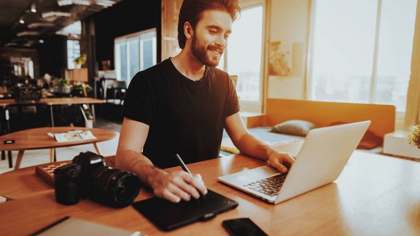 Smiling Graphic Designer Works on Laptop Indoors. Portrait of Bearded Stylish Professional Photographer Uses Digital Tablet Sits at Workplace Retouches Photo. Freelancing and Remote Work Concept - Фото, изображение