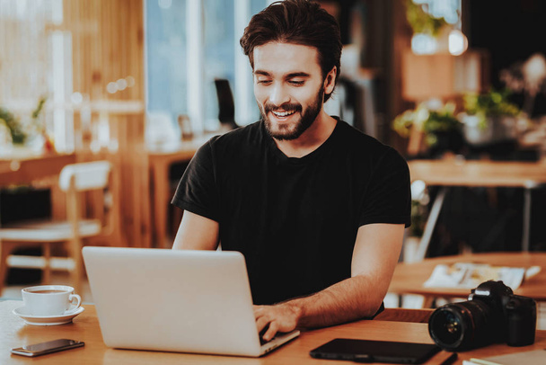 Portrait of Smiling Freelancer Working on Laptop. Handsome Caucasian Hipster Graphic Designer Wears Black T-Shirt Sits at Table with Gadgets Indoors. Distance Job and Freelancing Concept - Foto, Bild