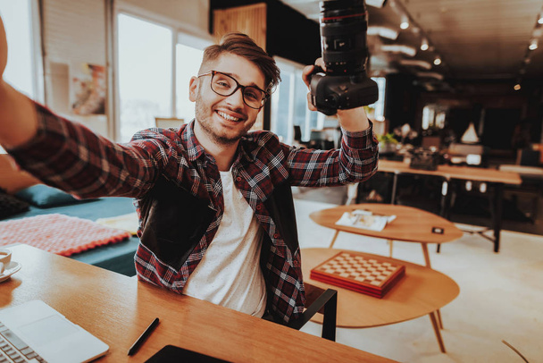 Smiling Guy Make Selfie when Working at Workplace. Portrait of Stylish Bearded Smiling Handsome Caucasian Photographer Wearing Glasses Holds Professional Camera in Hand Sits at Table Indoors. - 写真・画像