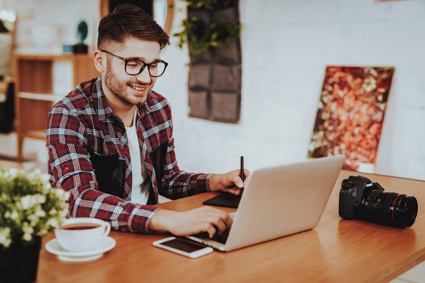 Portrait of Smiling Freelancer Working on Laptop. Handsome Caucasian Hipster Graphic Designer Wears Glasses Sits at Table Using Digital Tablet with Stylus Pen Indoors. Distance Job Concept - Foto, Bild