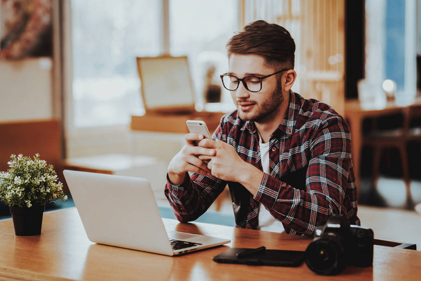 Freelancer Uses Phone and Work on Laptop Remotely. Portrait of Serious Stylish Graphic Designer Wears Glasses Sits at Table Indoors. Distance Job and Freelancing Communication Concept - Foto, Imagem