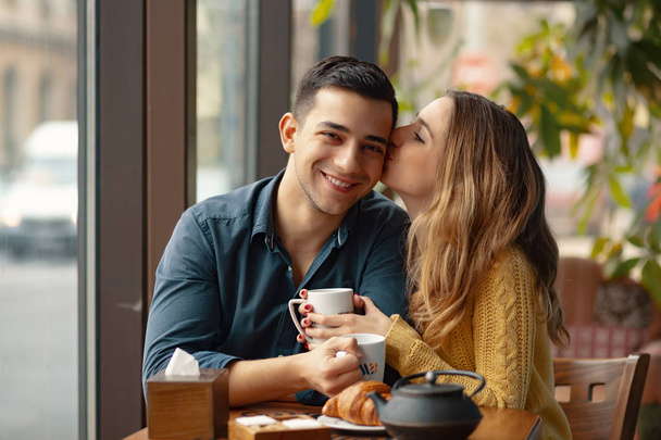 Mujer besando a sus hombres en la mejilla con afecto. Joven pareja atractiva en la fecha en la cafetería
.  - Foto, imagen
