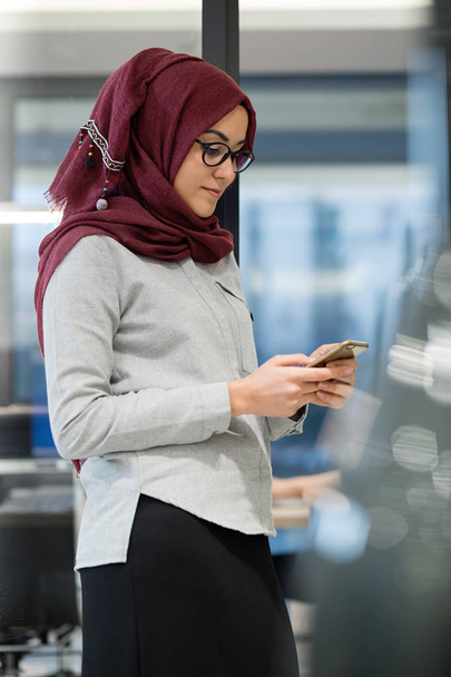 A young Muslim woman wearing a turban (hijab) is working on her phone by standing in office. She is elegant, beautiful and professional. - Photo, Image