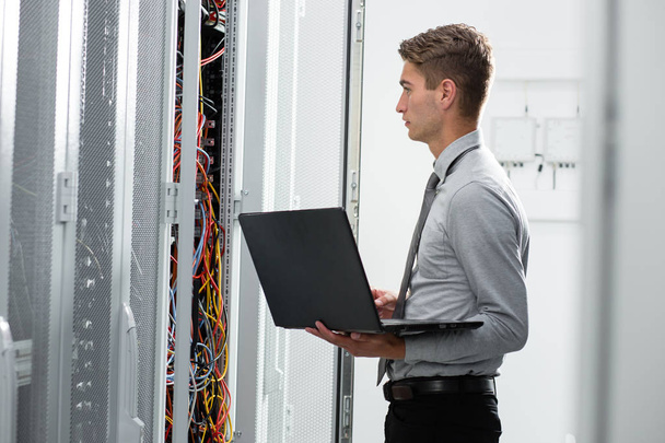 Male Server Engineer Works on a Laptop in Large Data Center. - 写真・画像