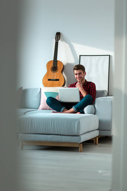 Young millennial positive man sitting on couch in living room with laptop alone on weekend at home - Foto, Bild