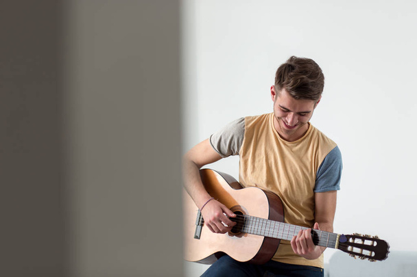 Pretty young man playing guitar while sitting on sofa in light living room - Fotografie, Obrázek