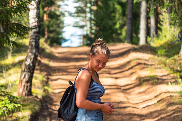 Jeune et belle fille marchant sur la route dans la forêt avec un sac sur les épaules lors d'une journée d'été ensoleillée avec un fond flou
 - Photo, image