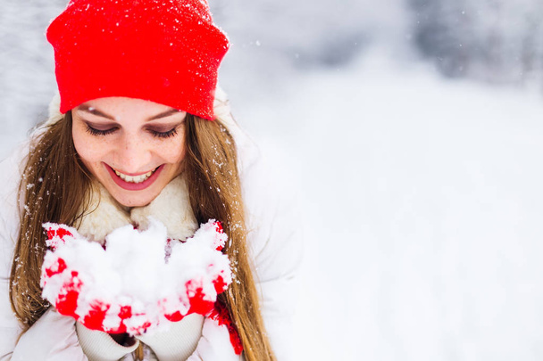 όμορφο χαμόγελο και μακιγιάζ το κορίτσι σε ένα χειμερινό μπουφάν και ζεστό αξεσουάρ που κρατά το χιόνι στα χέρια της - Φωτογραφία, εικόνα