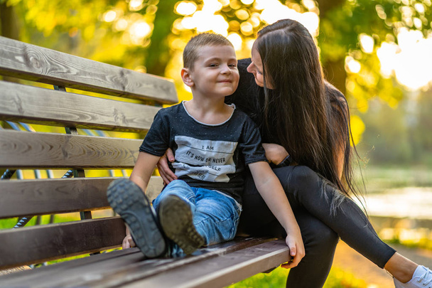 Niño dando besos a su madre en un banco del parque en Autum con fondo colorido en un día soleado - Leyenda en la camisa "Estoy, Es ahora o nunca, Me pregunto, ¿Por qué estoy aquí
" - Foto, imagen