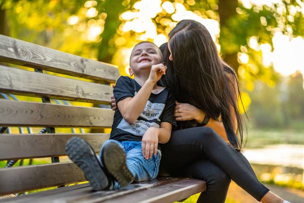 Mom Tickles Her Son on a Park Benck in Autum with Colorful Backgroun in a Sunny Day, Both Laughing- Legtion on Shirt "Eu sou, É agora ou nunca, Eu me pergunto, Por que eu estou aqui
" - Foto, Imagem