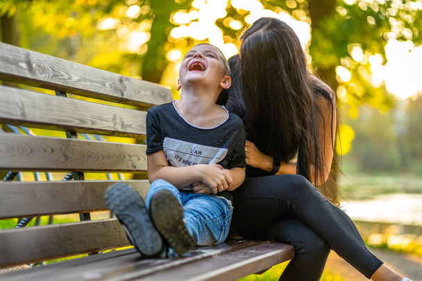 Mutter kitzelt ihren Sohn im Herbst auf einer Parkbank mit buntem Hintergrund an einem sonnigen Tag, beide lachen - Bildunterschrift auf Hemd: "Ich bin, es ist jetzt oder nie, ich frage mich, warum ich hier bin" - Foto, Bild