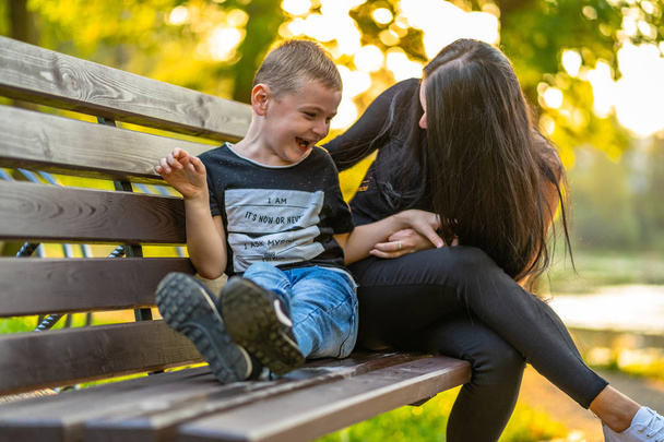 Mom Tickles Her Son on a Park Benck in Autum with Colorful Backgroun in a Sunny Day, Both Laughing- Legtion on Shirt "Eu sou, É agora ou nunca, Eu me pergunto, Por que eu estou aqui
" - Foto, Imagem