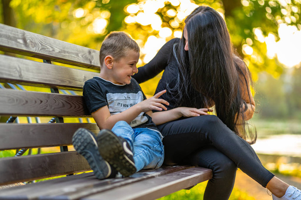 Mom Tickles Her Son on a Park Benck  in Autum with Colorful Backgroun in a Sunny Day, Both Laughing- Caption on Shirt "I am, It`s now or never, Iask myself, Why I`m here" - Valokuva, kuva