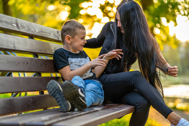 Mom Tickles Her Son on a Park Benck  in Autum with Colorful Backgroun in a Sunny Day, Both Laughing- Caption on Shirt "I am, It`s now or never, Iask myself, Why I`m here" - Photo, Image