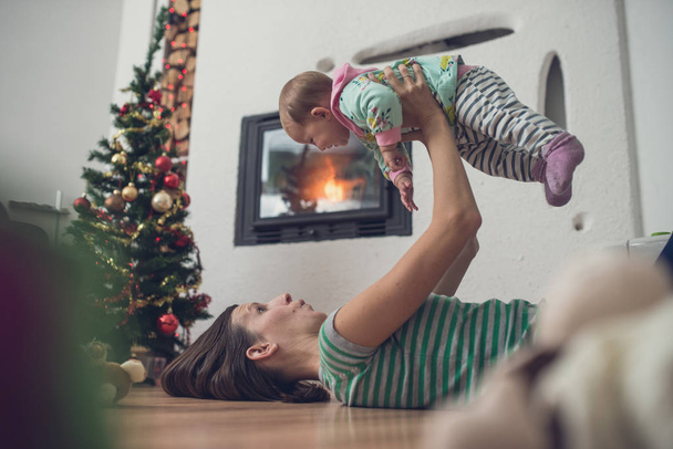 Retro image of playful young mother lying on the living room floor next to a fireplace and Christmas tree playing with her baby daughter lifting her high up in the air. - Foto, afbeelding