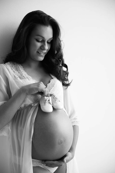 Jonge zwangere vrouw in lace Nachthemd houden babyslofjes op lichte achtergrond, zwart-wit effect. Ruimte voor tekst - Foto, afbeelding