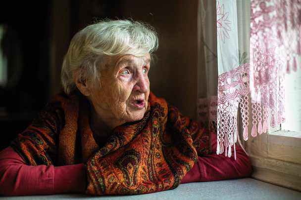 Porträt einer älteren Frau, die im Haus sitzt und aus dem Fenster schaut. - Foto, Bild