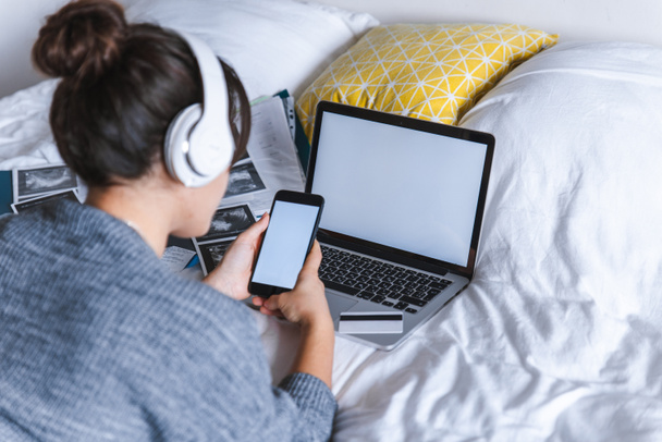 žena používající telefon v posteli laptop na pozadí. bílé prázdné obrazovky. kopie prostor - Fotografie, Obrázek