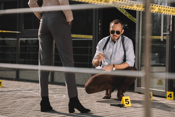 Sedící muž detektiv v sluneční brýle kouření a při pohledu na místo činu s partnerem stojící nablízku - Fotografie, Obrázek