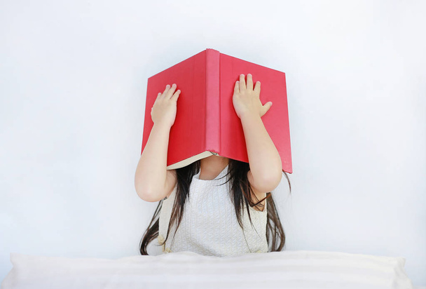 Little Asian child girl hiding face with open hardcover book sitting on bed with pillow. - Zdjęcie, obraz