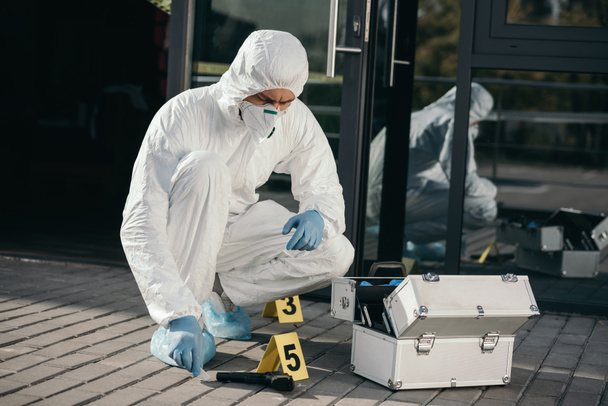 mannelijke criminoloog in beschermend pak en latex handschoenen zitten in de buurt van bewijs pistool - Foto, afbeelding