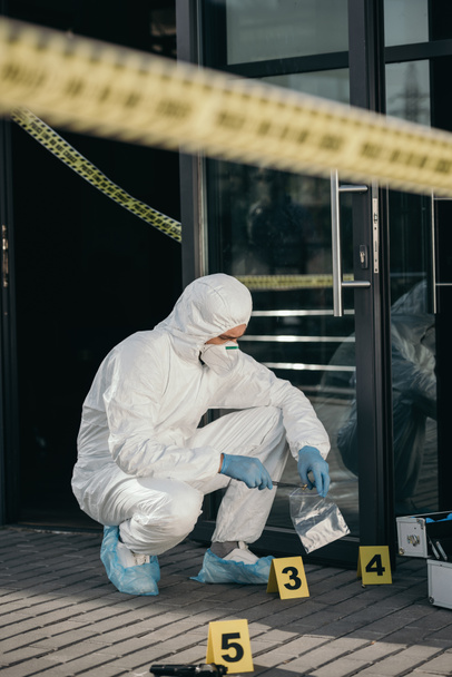 male criminologist in protective suit and latex gloves packing evidence with wizzles at crime scene - Fotoğraf, Görsel