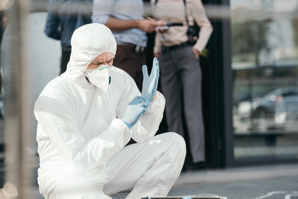 чоловічий кримінолог в захисному одязі латексних рукавичок з обрізаними детективами позаду
 - Фото, зображення