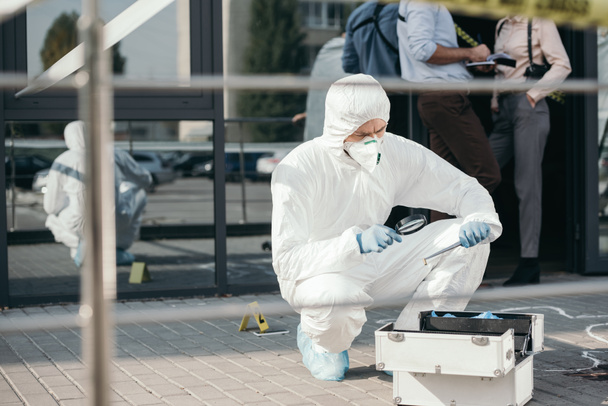 criminologista do sexo masculino em terno de proteção e luvas de látex olhando através de uma lupa em evidência
 - Foto, Imagem