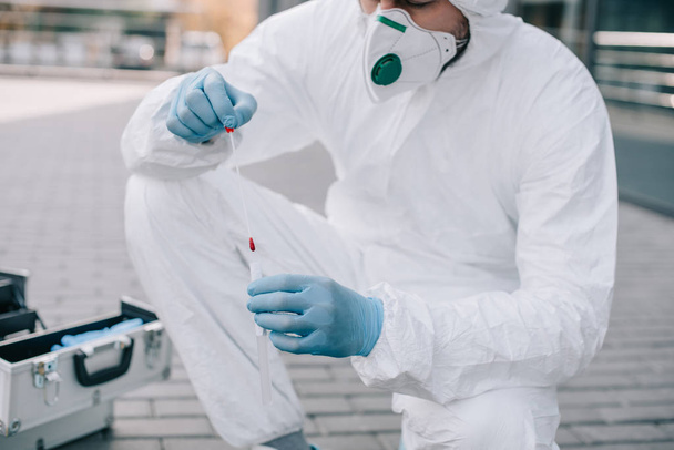 cropped view of male criminologist in protective suit and latex gloves putting evidence into test tube at crime scene - Photo, Image