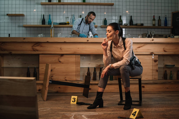 cropped view of smoking female detective sitting at crime scene with colleague working behind - Fotografie, Obrázek