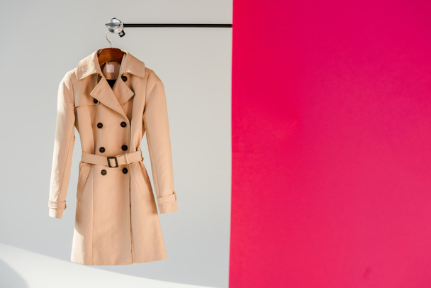 eleganter beiger Trenchcoat auf Kleiderbügel vor rosa und grauem Hintergrund  - Foto, Bild