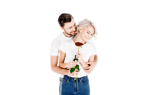 Όμορφο νεαρό ζευγάρι ενηλίκων αγκάλιασμα ενώ γυναίκα κρατώντας κόκκινο τριαντάφυλλο απομονωθεί σε λευκό  - Φωτογραφία, εικόνα