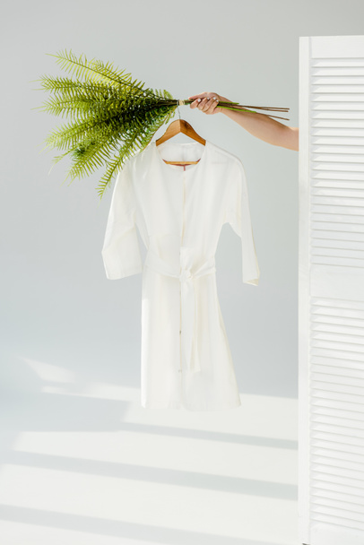 female hand holding hanger with white stylish dress and green plants - Foto, afbeelding