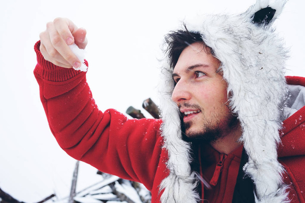  Jovem homem de casaco vermelho e chapéu peludo, aproveitando o dia de inverno nevado                               - Foto, Imagem