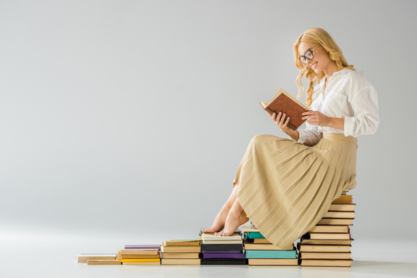 attractive smiling barefoot woman in glasses sitting on steps made of books   - Fotó, kép