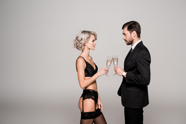 Atraktivní pár cinkání sklenic šampaňského, zatímco žena je v krásném prádle izolované Grey  - Fotografie, Obrázek