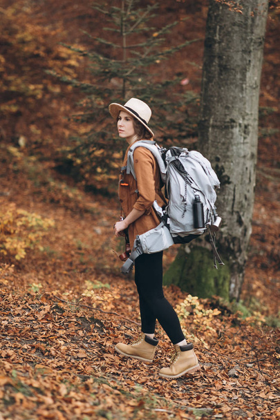 Young woman in a stylish hat and travel bag on her shoulders, looking around at the charming autumn forest - Foto, imagen