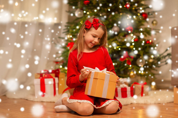 christmas, holidays and childhood concept - smiling girl in red dress with gift box at home - Foto, imagen