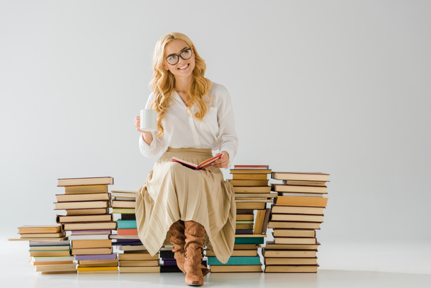 smiling beautiful woman drinking coffee and sitting on pile of books - Photo, Image