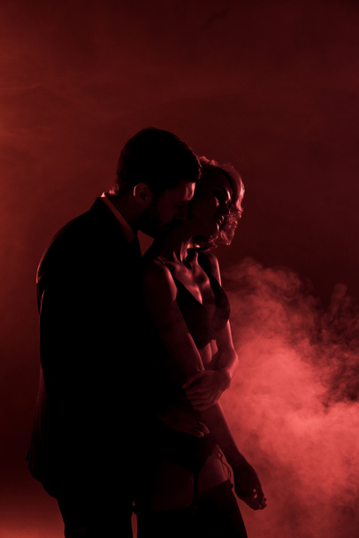 Man tenderly hugging beautiful woman and kissing her in neck on red smoke background - Foto, afbeelding