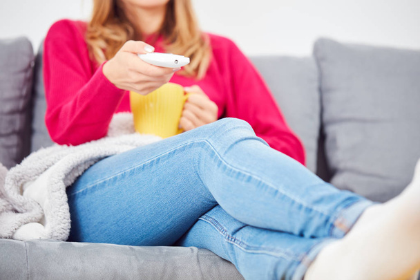 Κοπέλα που κρατά Tv remote και πίνοντας τον καφέ / τσάι σε έναν καναπέ. - Φωτογραφία, εικόνα