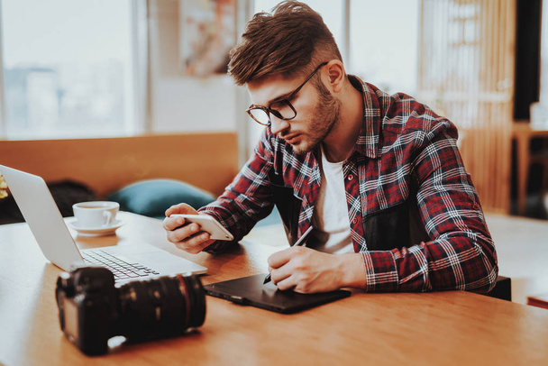 Freelancer Uses Smartphone and Working on Laptopp. Portrait of Handsome Caucasian Graphic Designer Wears Glasses Sits at Table Using Digital Tablet with Stylus Pen Indoors. Distance Job Concept - Фото, изображение
