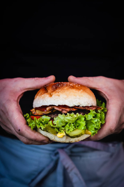 Male hands holding a juicy tasty cheeseburger with beef, lettuce, pickles, tomato and onion rings on a wooden table. Classic street food - grilled burger. - Photo, image