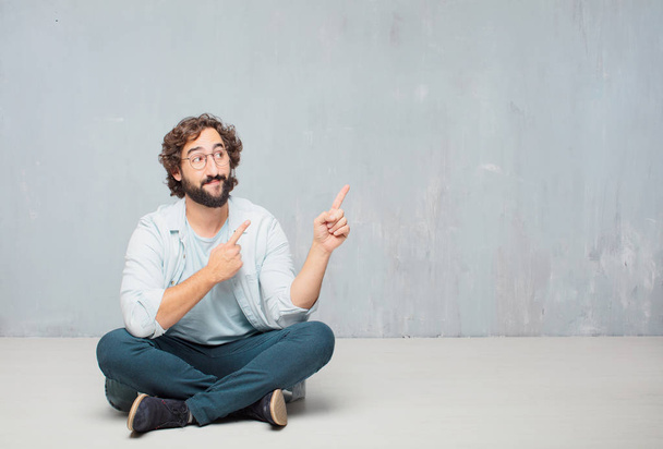 Young cool bearded man sitting on the floor. Grunge wall background. - Foto, Imagem