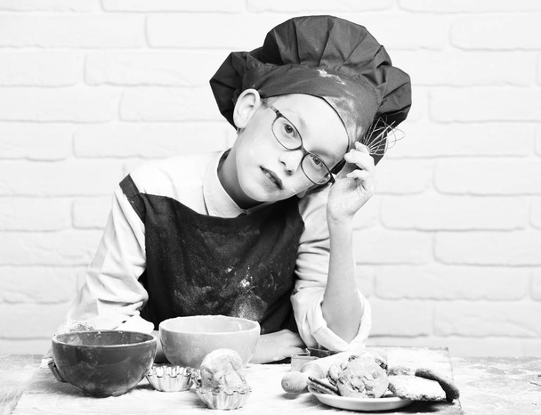 young boy cute cook chef in red uniform and hat on stained face with glasses sitting on table with colorful bowls, tasty cakes, rolling pin and kitchen whisk on white brick wall backgroun - Foto, immagini