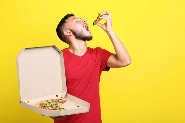 Νεαρός άνδρας τρώει πίτσα και κρατώντας κουτί από χαρτόνι σε κίτρινο φόντο - Φωτογραφία, εικόνα