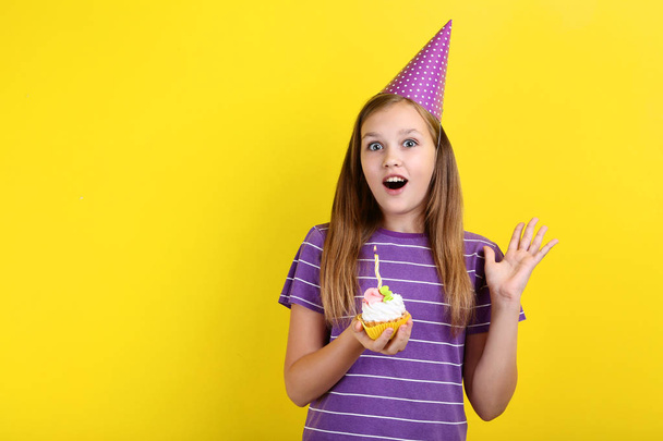 Young girl in birthday hat holding cupcake with candle on yellow background - Foto, Bild